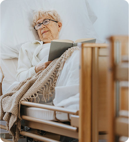 Una persona anziana in un letto per degenza, indossa occhiali e legge un libro, avvolta in una coperta beige.