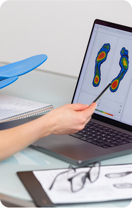 Un professionista mostra un'analisi di una scansione plantare su un computer portatile, evidenziando le diverse zone di pressione e supporto del piede. Sullo sfondo, si vedono delle solette blu e un quaderno.